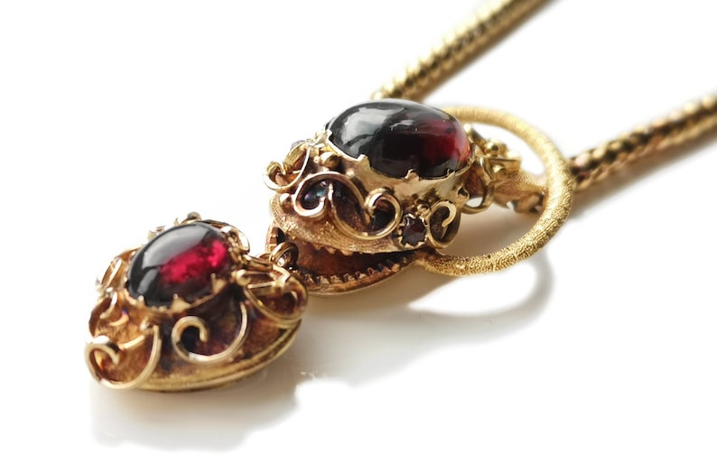May include: A gold necklace with a pendant featuring two red gemstones set in ornate gold frames. The pendant is attached to a delicate gold chain.