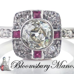 May include: A silver ring with a large, round, clear diamond surrounded by smaller diamonds and four red ruby accents. The ring has a square-shaped setting.