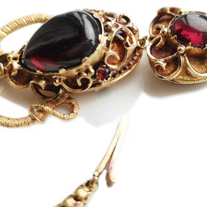May include: A gold necklace with a heart-shaped pendant featuring a red gemstone. The pendant is attached to a gold chain with a decorative clasp.