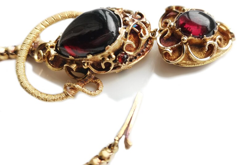 May include: A gold necklace with a heart-shaped pendant featuring a red gemstone. The pendant is attached to a gold chain with a decorative clasp.