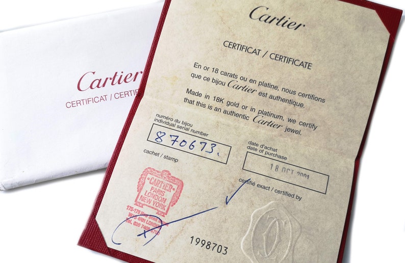 May include: A red Cartier certificate with the text 'Cartier Certificat/Certificate' in black. The certificate states that the jewelry is made of 18K gold or platinum and is authentic. The individual serial number is 870673 and the date of purchase is 18 Oct 2001. The certificate is stamped with a Cartier logo and the number 1998703.
