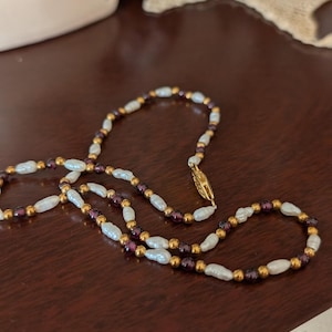 Peut inclure: Un collier délicat composé de perles blanches ovales, entrecoupées de petites perles dorées et de perles violettes foncées. Le collier est fermé par un fermoir doré et repose sur une surface brun foncé.