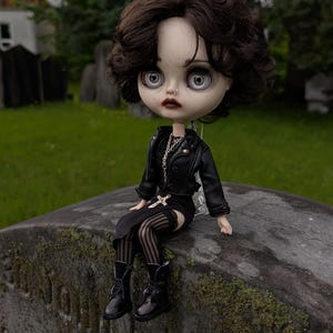 May include: A doll with short dark wavy hair, pale skin, and large grey eyes. The doll wears a black leather-style jacket, a black top, a black skirt, striped tights, and black boots. A silver chain necklace with a cross pendant is visible.