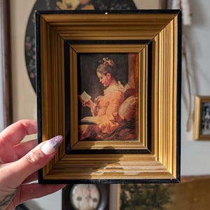 Peut inclure: Petite impression encadrée représentant une femme lisant. L'œuvre est dans un cadre doré et noir. La femme porte une robe couleur pêche. Le cadre a une finition dorée texturée.