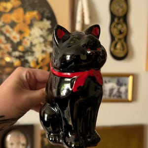 May include: A glossy black ceramic cat figurine in a seated position. The cat features a bright red scarf tied around its neck, with matching red accents inside its pointed ears. The surface is smooth and reflective, highlighting the feline form.
