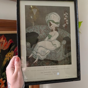 Peut inclure: Une impression vintage encadrée intitulée A MAN-TRAP représentant une personne en robe blanche et grand bonnet du XVIIIe siècle assise sur un canapé à motifs. L'impression comporte du texte en anglais et en allemand, dans un cadre noir.