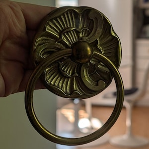Peut inclure: Poignée de porte en laiton orné avec une poignée circulaire. La poignée présente un motif floral avec des sculptures détaillées et une sphère centrale. La poignée est fixée à une base circulaire décorative.