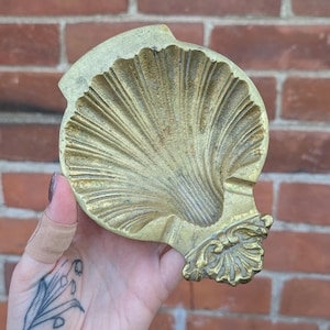 Peut inclure: Un plat en laiton doré en forme de coquillage présentant une surface intérieure festonnée avec des crêtes rayonnantes et un élément de base ornementé. L'objet a une forme de bol large et ouvert avec une finition métallique texturée.