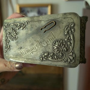 Peut inclure: Une boîte rectangulaire en métal argenté terni avec des motifs de volutes en relief aux coins. Le couvercle porte l'inscription cursive "Woman's Friend" et comporte une petite épingle à cheveux en métal en forme de U fixée dessus.