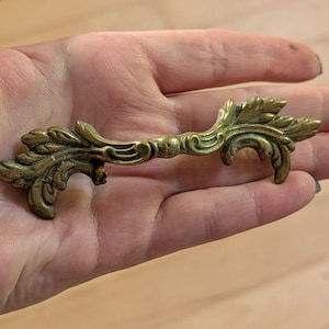 Peut inclure: Poignée de tiroir en laiton ornée, avec un design symétrique détaillé. La quincaillerie présente une section de poignée centrale avec des éléments décoratifs de feuilles et de volutes à chaque extrémité. La finition vieillie suggère une esthétique vintage, pour meubles.