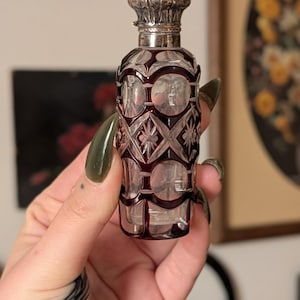 May include: An antique perfume bottle with a silver cap and a cut glass body. The glass is clear with a deep red overlay, featuring geometric and floral patterns. The bottle is held up against a neutral background.