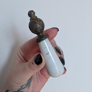 Peut inclure: Poignée de meuble antique en céramique blanche et bronze. La poignée a un corps blanc en forme de goutte d'eau avec un dessus en bronze. Le dessus en bronze a un design arrondi décoratif. Une pièce de quincaillerie vintage.