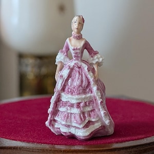 以下が含まれることがあります： ピンクと白のガウンを着た女性の磁器製フィギュア。フリル袖とハイカラーが特徴です。フィギュアは、赤いベルベットの天板が付いた小さな丸いテーブルの上にあります。歴史的な人物のミニチュア表現です。