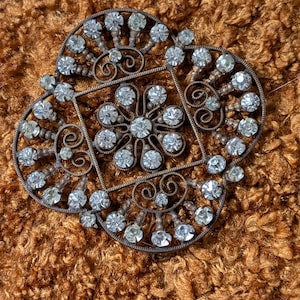 Peut inclure: Broche ancienne en forme de trèfle à quatre feuilles. La broche présente des strass clairs et étincelants sertis dans un cadre métallique orné et détaillé. Le métal a une patine chaude et vieillie. La broche est posée sur une surface brune texturée.