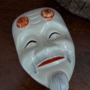 Op de afbeelding: Een wit keramieken masker met een gestileerd gezicht met gesloten ogen, een rode mond en een grijze sik. Twee oranje cirkelvormige patronen zijn op het voorhoofd geschilderd. Het masker heeft een glanzende afwerking en een ovale vorm.