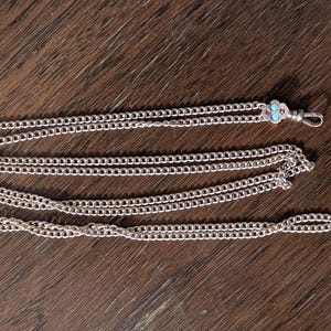 May include: Silver-toned chain necklace with a decorative clasp. The necklace is composed of multiple strands of small, linked chains. The clasp features a floral design with small, turquoise-colored accents. The necklace is displayed on a wooden surface.