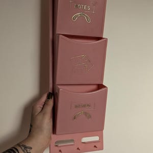 May include: A pink plastic wall organiser with three compartments for notes, mail, and memos. Each compartment has a gold inscription with a decorative design.