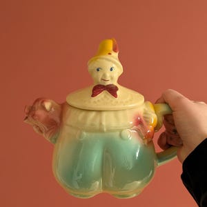 Puede incluir: Una tetera de cerámica vintage con un diseño caprichoso. La tetera presenta una figura en la tapa con un sombrero amarillo y una pajarita roja. El cuerpo de la tetera es de color verde claro y amarillo, con una boquilla en forma de cerdo. El asa es amarilla.