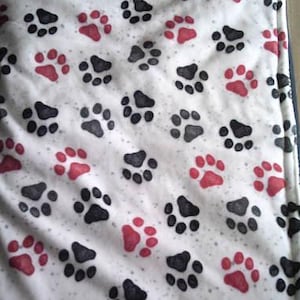 May include: White pet bed with a pattern of black and red paw prints. The bed has a dark blue border and is rectangular in shape. The paw prints are evenly spaced across the surface of the bed.