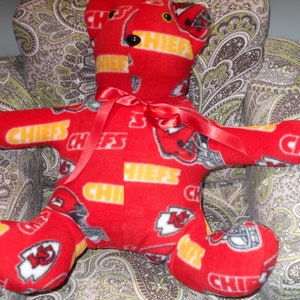 May include: Red plush teddy bear with a Kansas City Chiefs pattern. The bear is wearing a red ribbon around its neck and is sitting in a chair with a paisley pattern.
