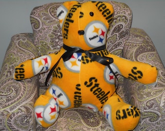 May include: A yellow plush teddy bear with the Pittsburgh Steelers logo and the word "Steelers" repeated on the fabric. The bear is wearing a black ribbon around its neck and is sitting on a patterned armchair.