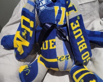 May include: A blue and yellow plush bunny with a black nose and blue eyes. The bunny is made from fabric scraps with the words "Go Blue" and "Blue" on it. The bunny has a blue ribbon tied around its neck.