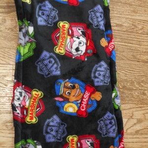 May include: Black fleece blanket with a repeating pattern of Paw Patrol characters, including Chase and Marshall. The characters are wearing their signature uniforms and have their names printed on their badges.