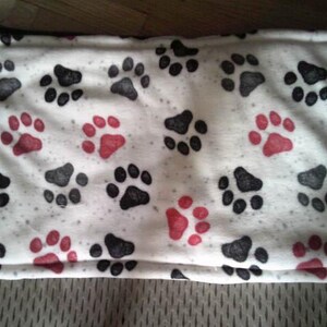 May include: A rectangular pet bed with a white background, covered in a pattern of black and red paw prints. The bed has a soft, plush texture and a white border. Ideal for a dog or cat.