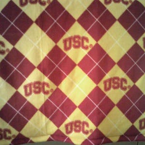 May include: A fabric with a repeating pattern of red and yellow diamonds, with white lines separating them. The letters "USC" are printed in red within the yellow diamonds. The overall design is reminiscent of a classic argyle pattern.