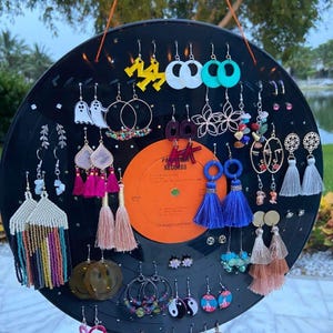 Può includere: Un disco in vinile nero con una varietà di orecchini esposti su di esso. Gli orecchini sono realizzati con diversi materiali, tra cui metallo, plastica e perline. Alcuni degli orecchini hanno la forma di fantasmi, fiori e facce sorridenti. Il disco è appeso a una corda.