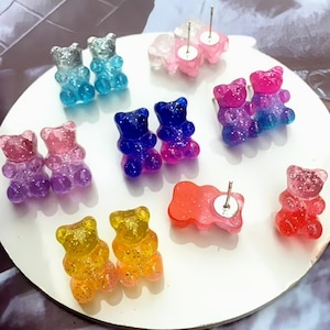 Op de afbeelding: Een verzameling gummy bear oorknopjes in verschillende kleuren, waaronder blauw, roze, paars, geel en rood. De oorbellen zijn gerangschikt op een wit oppervlak, met glitteraccenten op sommige paren.