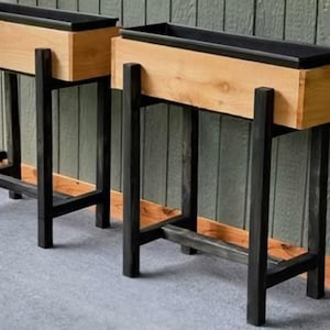 Puede incluir: Dos jardineras rectangulares de madera con patas de metal negro. Las jardineras tienen un acabado de madera natural con interior negro. Las patas son de un negro oscuro mate, y el diseño es moderno y minimalista. Adecuado para uso en interiores o exteriores.