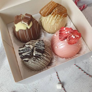 May include: A box containing four chocolate spheres, each with a unique decoration. One is dark chocolate with white drizzle and a Kinder Bueno. Another is light-coloured with white drizzle and a biscuit. The third is pink with red accents. The fourth is grey with Oreo pieces.
