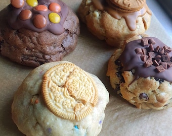 May include: Four large, freshly baked cookies on parchment paper. One cookie is topped with chocolate frosting and colorful candies. Another has a cookie on top with caramel drizzle. The third has an Oreo cookie on top, and the last has chocolate chips and chocolate frosting.