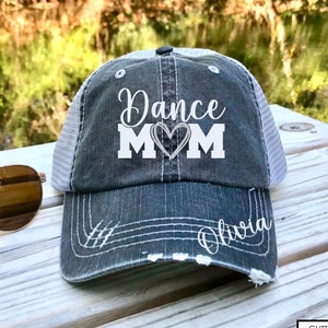May include: A gray and white distressed baseball cap with the text "Dance Mom" and a heart design. The name "Olivia" is embroidered on the back of the cap.