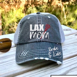 May include: A gray and white distressed baseball cap with the text "LAX MOM" embroidered in white with a red lacrosse stick. The text "Drake & Luke" is embroidered in white on the back of the cap.