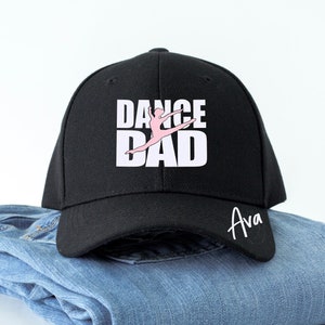 May include: Black baseball cap with white text that reads "Dance Dad" and a pink silhouette of a dancer. The cap has a black brim and a white name "Ava" written on the back.