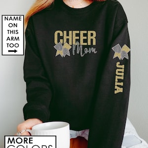 Może przedstawiać: Czarny sweter z napisem "Cheer Mom" z błyszczących złotych liter i złotą błyszczącą kokardką po obu stronach. Na prawym rękawie znajduje się złota błyszcząca kokardka i imię "Julia" napisane pionowo.
