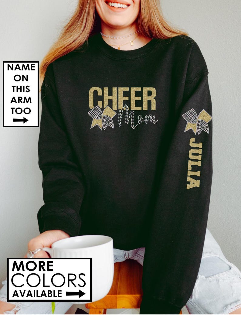 Może przedstawiać: Czarny sweter z napisem "Cheer Mom" z błyszczących złotych liter i złotą błyszczącą kokardką po obu stronach. Na prawym rękawie znajduje się złota błyszcząca kokardka i imię "Julia" napisane pionowo.