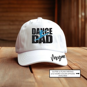 May include: White baseball cap with black glitter text that reads "Dance Dad" with a blue silhouette of a dancer. The cap has a black brim and the name "Megan" is written in black cursive on the back.