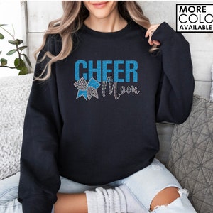 Może przedstawiać: Czarna bluza z napisem "Cheer Mom" w niebieskich literach z brokatem i niebieską i białą kokardką z brokatem.