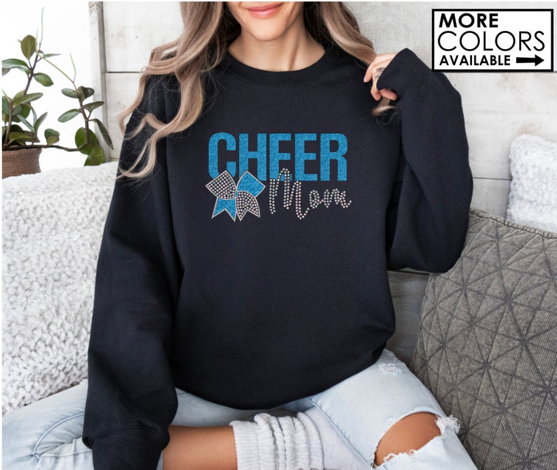 Może przedstawiać: Czarna bluza z napisem "Cheer Mom" w niebieskich literach z brokatem i niebieską i białą kokardką z brokatem.