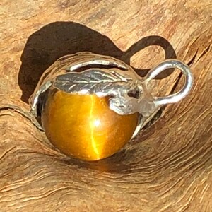 Könnte beinhalten: Silberanhänger mit einem Tigerauge-Edelstein. Der goldbraune Edelstein ist in eine silberne Fassung in Blattform eingefasst. Der Anhänger hat eine kleine Schlaufe oben für eine Kette.