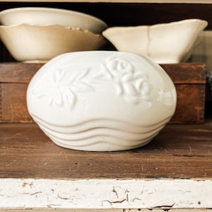 May include: White ceramic planter with a raised floral design and wavy edges. The planter is sitting on a wooden shelf.