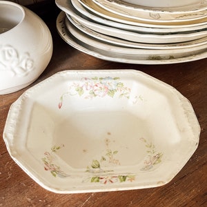 May include: A white ceramic serving bowl with a floral design in shades of pink, blue, and green. The bowl has an octagonal shape and a slightly raised rim.