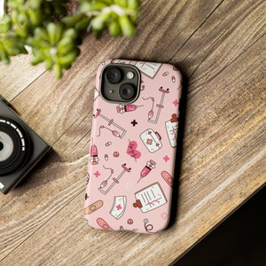 Puede incluir: Funda de teléfono rosa con un patrón de ilustraciones médicas, incluyendo jeringas, estetoscopios y vendajes. El diseño incluye un gráfico de latido cardíaco. Colocada sobre una superficie de madera.