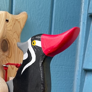 Könnte beinhalten: Eine hölzerne Specht-Wanddekoration mit rotem Schnabel und schwarz-weißen Federn. Der Specht ist an einer Holzbasis mit einer roten Schnur befestigt, an der unten eine rote Kugel hängt.