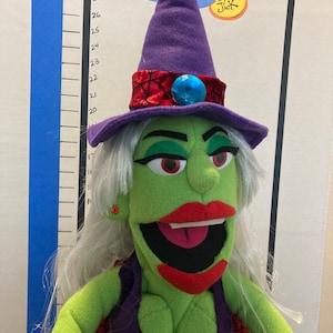 May include: A green witch puppet with a purple pointed hat, red lips, and white hair. The puppet has a purple dress and a red spiderweb cape. The puppet is in front of a measuring chart and a sign that says "Dramatic Puppets by Jack."