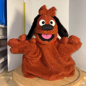May include: A brown plush dog puppet with black ears, white eyes, and a pink tongue. The puppet has outstretched arms and is sitting on a wooden cutting board. The puppet has a red collar.
