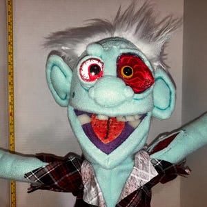 May include: A plush puppet with a light blue face, large ears, and white hair. It has one normal eye and one red, bloodshot eye. The puppet wears a plaid shirt and has outstretched arms.